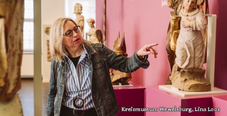 Auf dem Bild ist eine Dame zu sehen, die mit der Hand auf ein Ausstellungsstück zeigt. Zu sehen ist eine Figur.